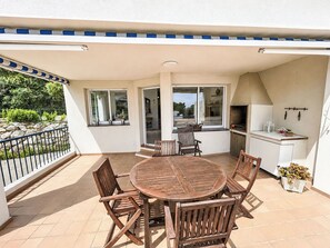 Une terrasse couverte avec une table et des chaises en bois rondes, un barbecue et un petit coin cuisine.