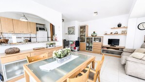 A modern kitchen with wooden cabinets, a dining table with chairs, a living area with a sofa, and a television. - El Jardinet by Interhome (Llançà)