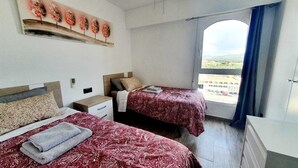 Une chambre d’hôtel avec deux lits, une petite table de chevet, une commode, une fenêtre donnant sur un beau paysage et une œuvre d’art accrochée au mur.
