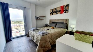 Une chambre d’hôtel avec un grand lit, une table de chevet, une commode, une fenêtre avec des rideaux, une vue sur l’extérieur et une œuvre d’art murale.