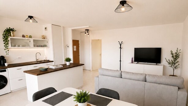 Et moderne køkken med hvide skabe, et spisebord med fire stole, en grå sofa, et fjernsyn på et stativ og en plante i vindueskarmen.