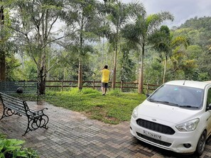 Un chemin pavĂ© avec un banc, une voiture blanche et une personne debout prĂšs dâune clĂŽture, dans un parc arborĂ©.