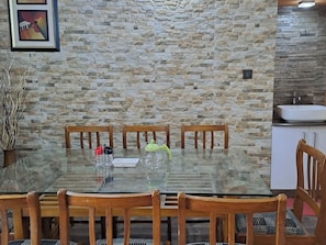 A dining area with a glass table, wooden chairs, a stone wall, and a kitchen area with a sink.