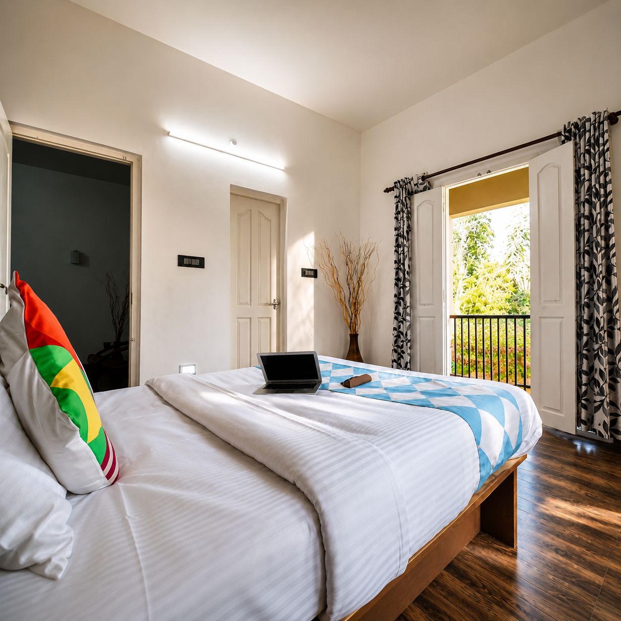 A bedroom with a bed, a laptop, a door, a window with curtains, and a view of greenery outside.