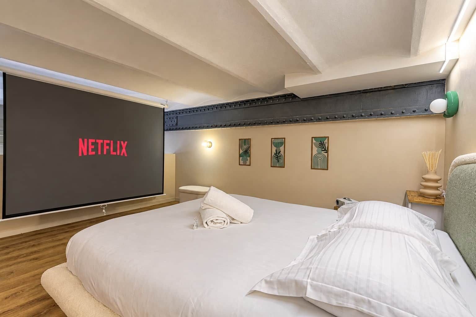 A hotel room with a bed, a wall-mounted TV displaying 'NETFLIX', a bedside table with a lamp, and a wall with framed artwork.