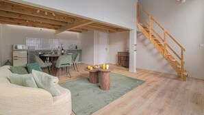 Een ruime woonkamer met een houten trap, een eettafel met stoelen, en een keuken op de achtergrond.