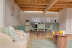 Un salon avec un canapé, des fauteuils verts, une table basse en bois et, au fond, une cuisine.