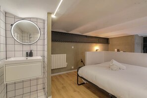 Habitación de hotel con una cama blanca, un espejo redondo, un lavamanos blanco, un radiador y suelo de madera.