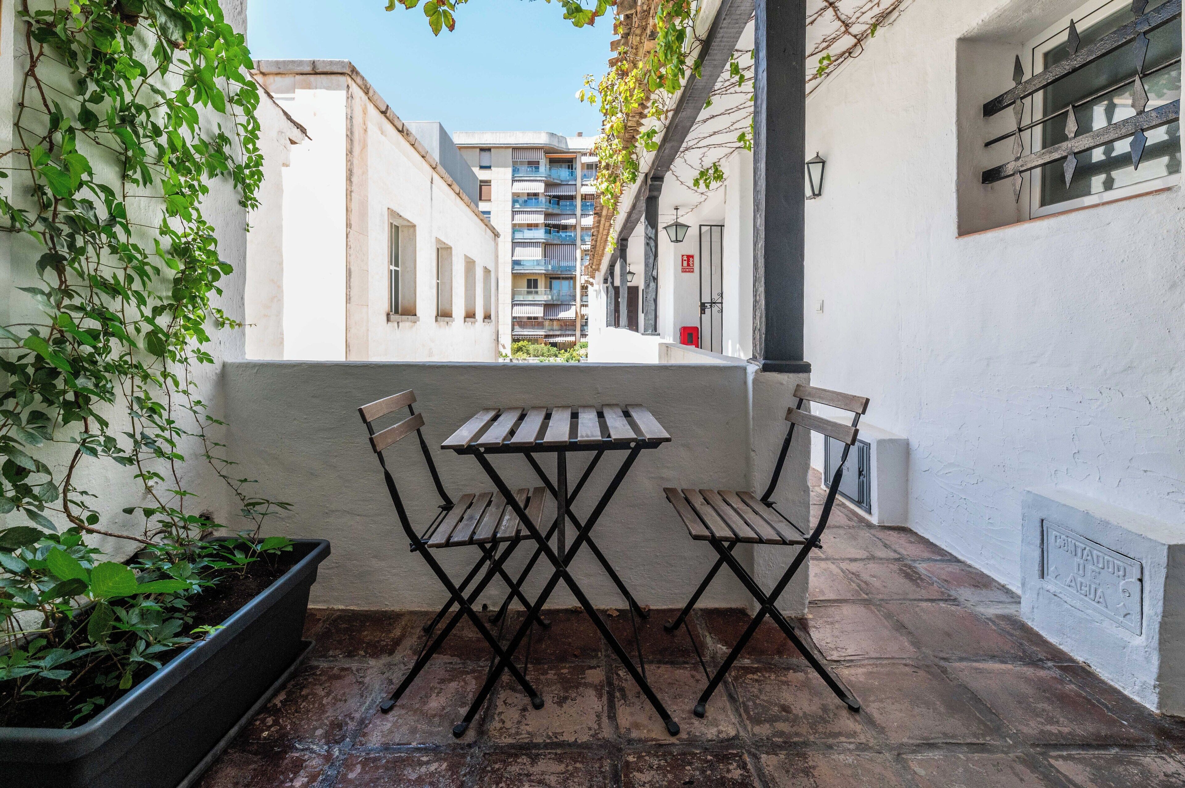 Een smal terras buiten, met een tafel en twee stoelen, omgeven door witte muren en een plant in pot.