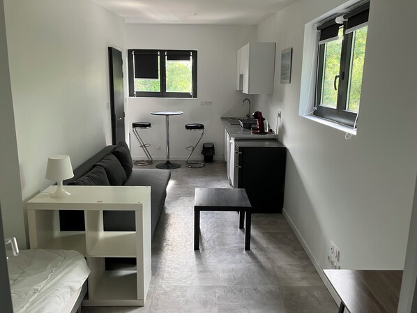 A compact living space with a sofa, a small table, a kitchenette, and a window with a view of greenery. - BRAND-NEW, FULLY EQUIPPED STUDIO IDEALLY LOCATED NEAR LA TORCHE (Plomeur)