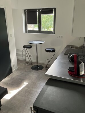 A kitchen with a window, two stools, a small table, and a red appliance. - BRAND-NEW, FULLY EQUIPPED STUDIO IDEALLY LOCATED NEAR LA TORCHE (Plomeur)