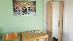 A room with a wooden chair, a small table with a magazine, a wooden wardrobe, and a painting of flowers on the wall. - Berghotel (Bad Oeynhausen)