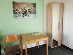 A room with a wooden chair, a small table with a magazine, a wooden wardrobe, and a painting of flowers on the wall. - Berghotel (Bad Oeynhausen)