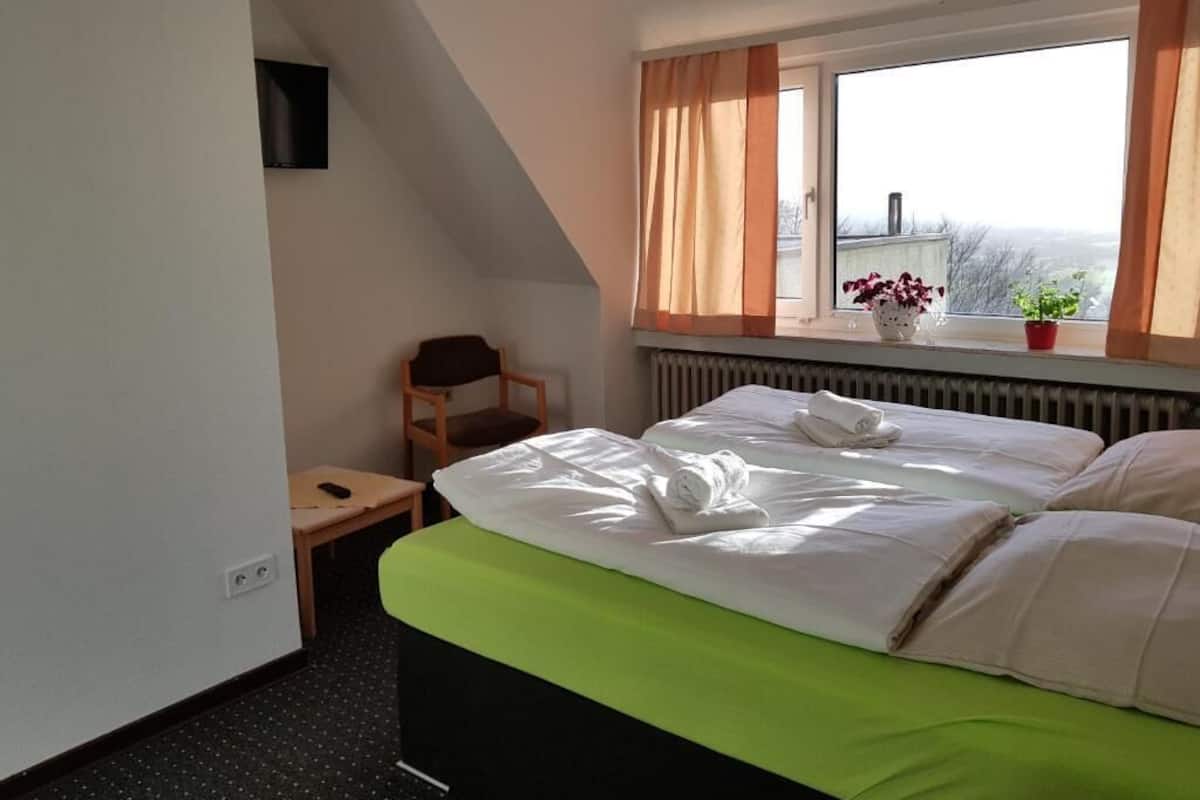 Una habitación de hotel con cama, una silla, una mesita, una ventana con cortinas y un radiador.