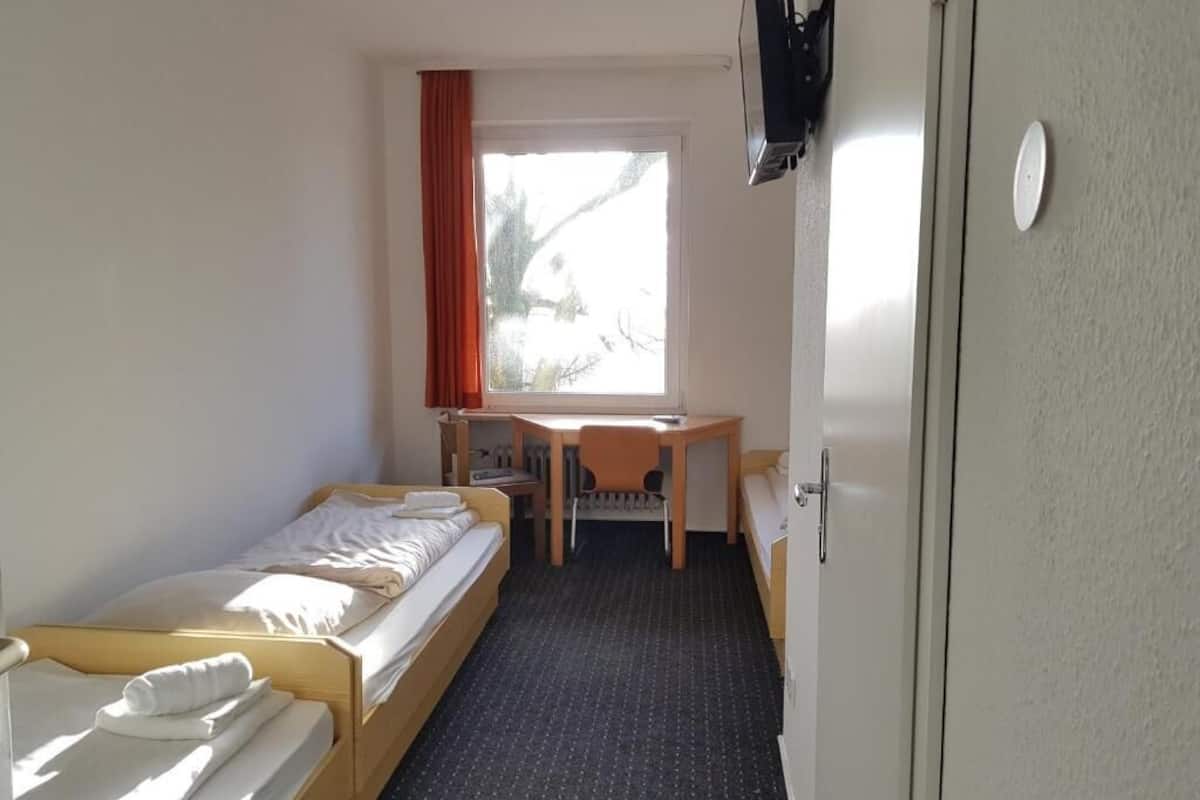 Una habitación con dos camas individuales, un escritorio pequeño, una silla, una ventana con cortinas y una puerta.