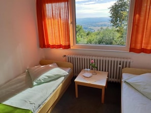 Ein kleines Zimmer mit zwei Einzelbetten, einem Tisch mit einer Vase und einer Blume sowie einem Fenster mit Vorhängen.