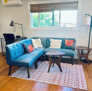 Ein Wohnzimmer mit einem blauen Ecksofa, einem kleinen runden Tisch, einer Lampe und einem Fenster mit Jalousien.