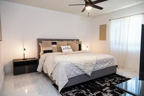 Un dormitorio moderno con una cama, mesitas de noche, un ventilador de techo y un amplio ventanal con cortinas.