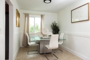 Uma sala de jantar com mesa de vidro, cadeiras brancas, um objeto decorativo na parede e uma janela com cortinas.