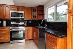 Una cocina con armarios de madera, un horno de acero inoxidable, un lavavajillas y una ventana con vista a palmeras.
