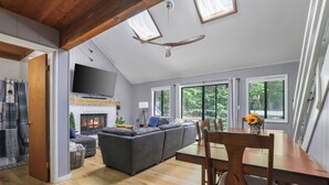 Ein geräumiger Wohnbereich mit Kamin, einem großen grauen Sofa, einem Esstisch mit Stühlen und einem an der Wand befestigten Fernseher.