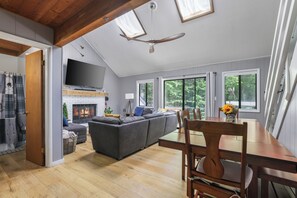 A spacious living area with a fireplace, a large gray sofa, a dining table with chairs, and a television mounted on the wall. - Little Bear Hideout Sauna, Game Room & Firepit (Lake Ariel)
