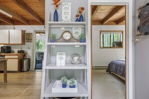 A room with a bed, a shelf with decorative items, a clock, and a window with a view of trees. - Little Bear Hideout Sauna, Game Room & Firepit (Lake Ariel)
