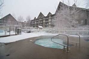 Area di una piscina scoperta ricoperta di neve con una vasca idromassaggio, circondata da edifici dallo stile architettonico tradizionale.