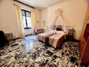A bedroom with a bed, a chair, a table, a window with curtains, and a decorative canopy.