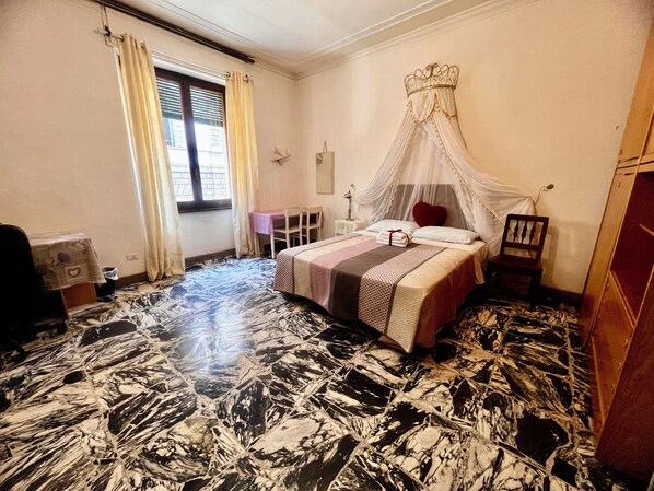 A bedroom with a bed, a chair, a table, a window with curtains, and a decorative canopy.