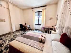 A hotel room with a bed, a desk, a chair, a small table, a potted plant, and a window with curtains.