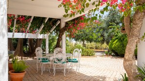 A covered patio with a table and chairs, surrounded by potted plants and trees.