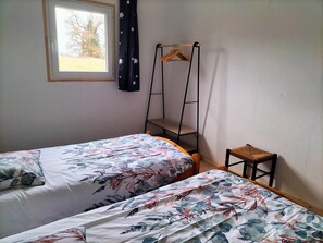 Ein Schlafzimmer mit zwei Betten, einem Fenster mit Blick auf BĂ€ume, einem hölzernen KleiderbĂŒgel und einem kleinen Hocker.