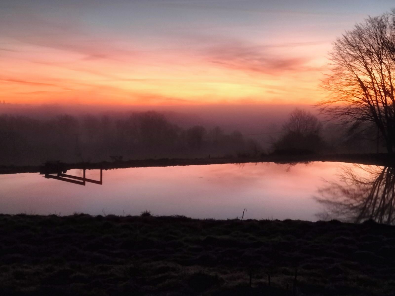 A serene landscape with a calm body of water, a wooden structure, and a tree silhouetted against a colorful sky.. Small wood-frame house for 4 people, quiet and comfortable.