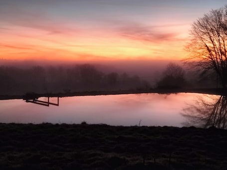 A serene landscape with a calm body of water, a wooden structure, and a tree silhouetted against a colorful sky.. Small wood-frame house for 4 people, quiet and comfortable.