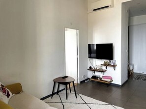 Ein Wohnzimmer mit einer Couch, einem kleinen Tisch, einem an der Wand befestigten Fernseher und einer Tür zu einem anderen Raum.