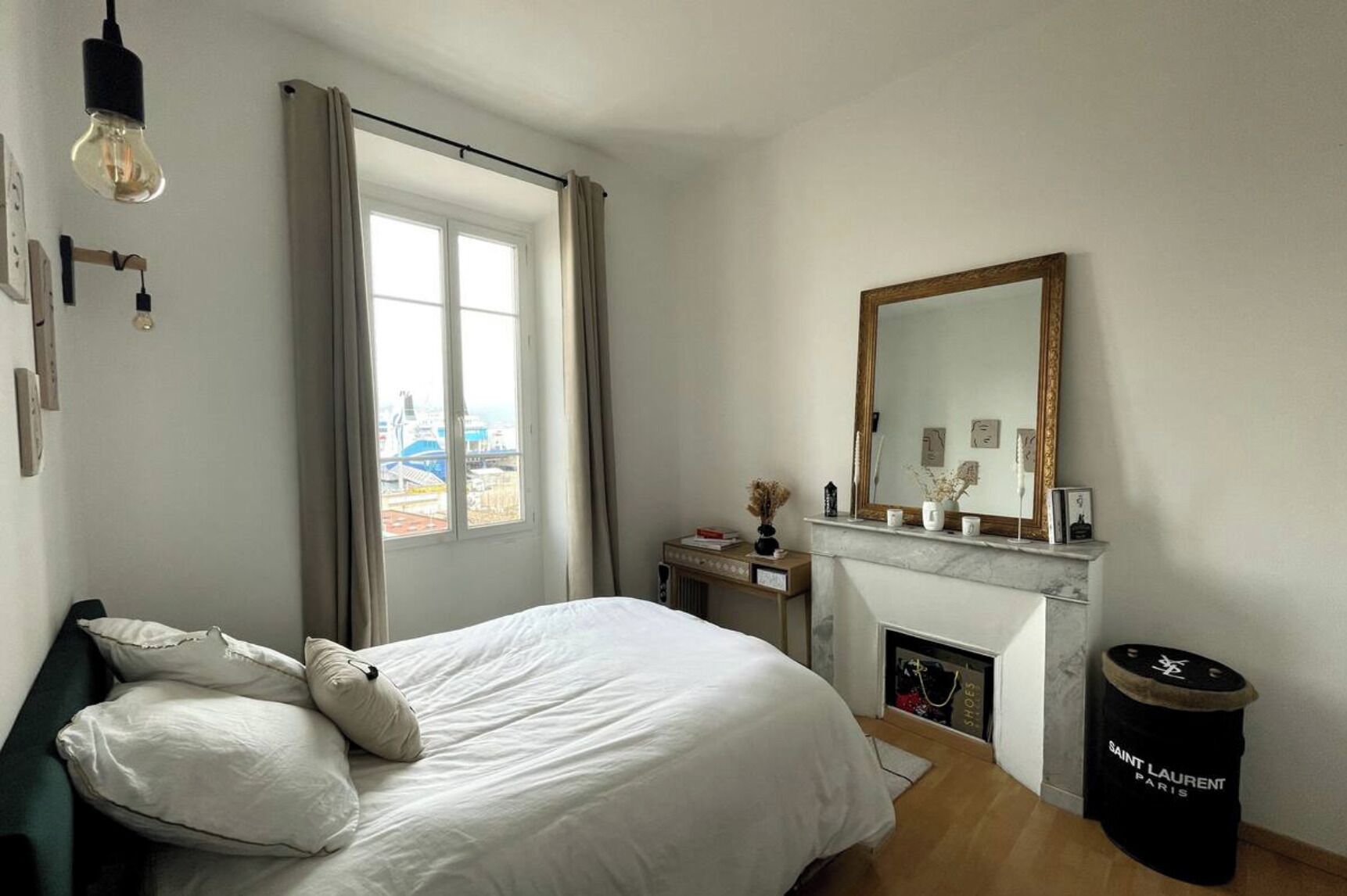 A bedroom with a bed, a desk, a fireplace, a large window, and a mirror.