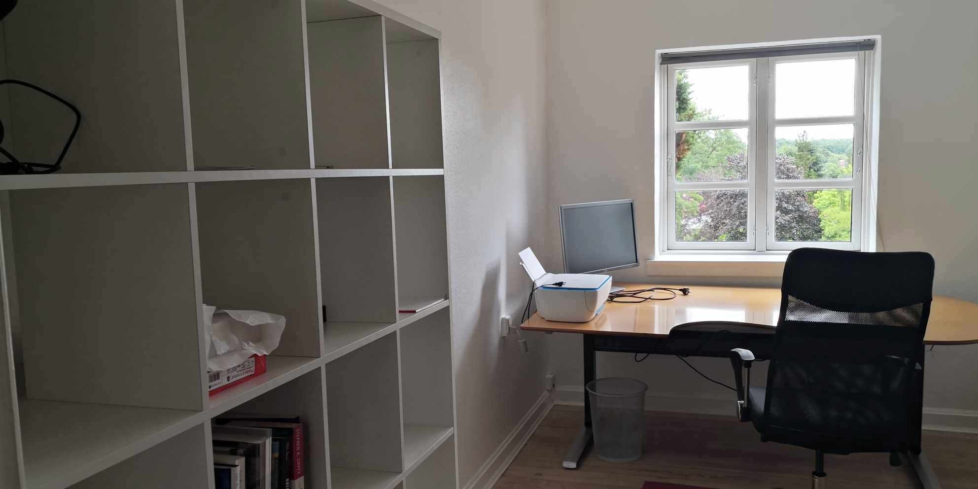 A room with a desk, chair, computer, bookshelf, and window with a view.