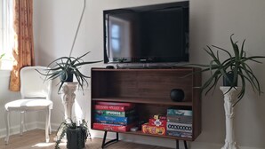 Een woonkamer met een televisie, een witte stoel, een houten kast met gezelschapsspellen en twee potplanten.