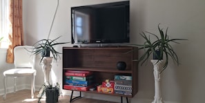Una sala de estar con una televisión, una silla blanca, un mueble de madera con juegos de mesa y dos plantas en macetas.