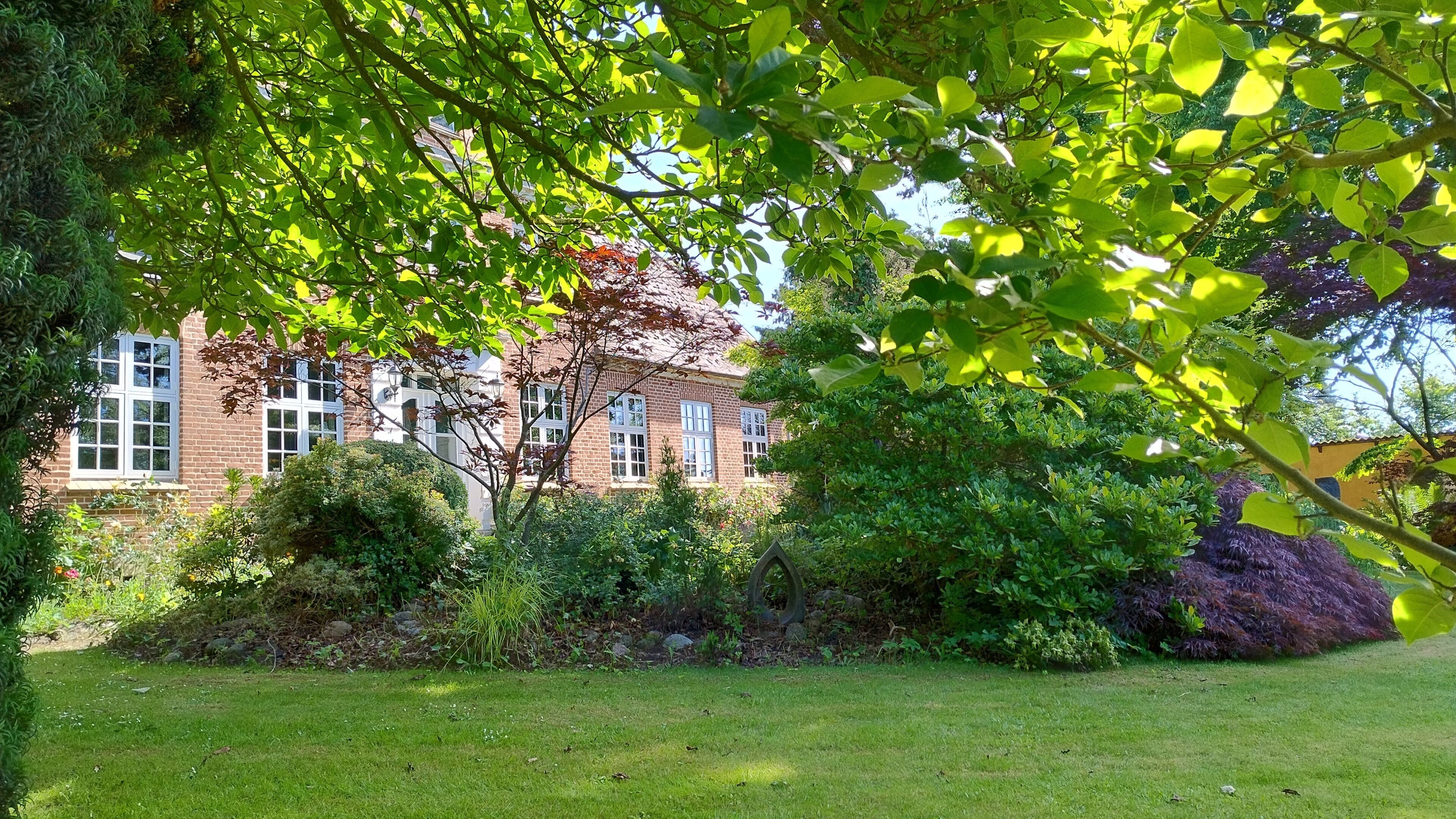 A brick building with multiple windows, surrounded by a well-maintained garden with various plants and a lawn.
