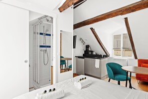 Ein modernes Zimmer mit Dusche, einem Bett mit Handtüchern, einem Essbereich mit Tisch und Stühlen sowie einer Küchenzeile.