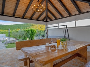 A wooden table with a bench, a chandelier, and a view of a green landscape. - Three Bedroom Holiday Home with Private Pool and Terrace (Šibenik)