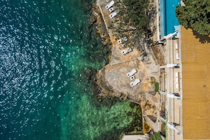 Luftaufnahme einer Küstenlandschaft mit klarem, blauem Wasser, einem Sandstrand, Liegestühlen und einem Gebäude mit einem Balkon mit Blick aufs Meer.