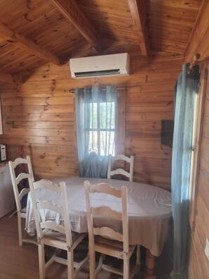 A wooden cabin with a dining table, four chairs, an air conditioning unit, and a window with curtains.