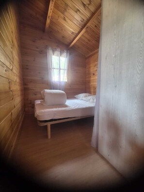 A small, wooden cabin room with a single bed, a window with curtains, and a wooden ceiling.
