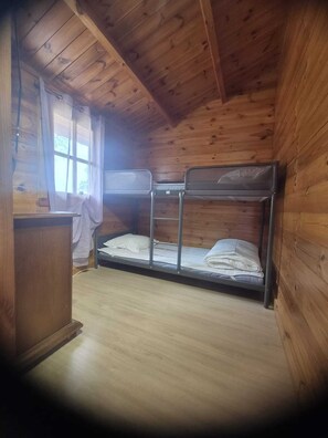 A wooden cabin room with bunk beds, a small desk, and a window with curtains.