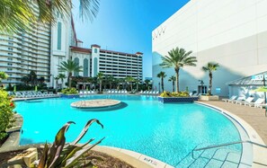 A hotel pool area with a large pool, lounge chairs, palm trees, and a modern building in the background. - OCEAN VIEW - 8th Floor, Sleeps 8, 2 bedrooms, 2 bathrooms and a bunk room. (Panama City Beach)