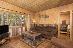 Ein Zimmer mit Holzwänden, einem großen Fenster, einem braunen Ledersofa, einem Holztisch und einem Fernseher auf einem Standfuß.