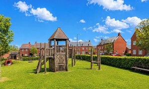 Ein Spielplatz mit einer Holzspielanlage, umgeben von einem gepflegten Rasen und Wohnhäusern im Hintergrund.
